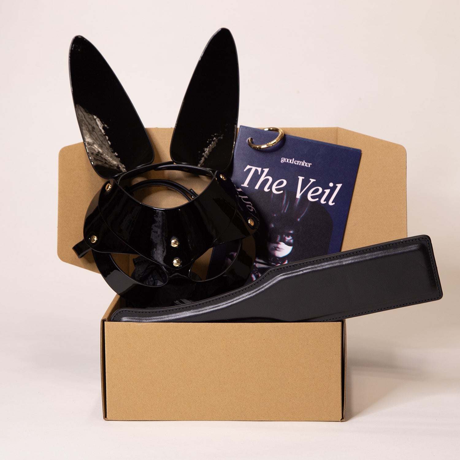 Open box containing a black bunny mask, a leather paddle and a booklet.
