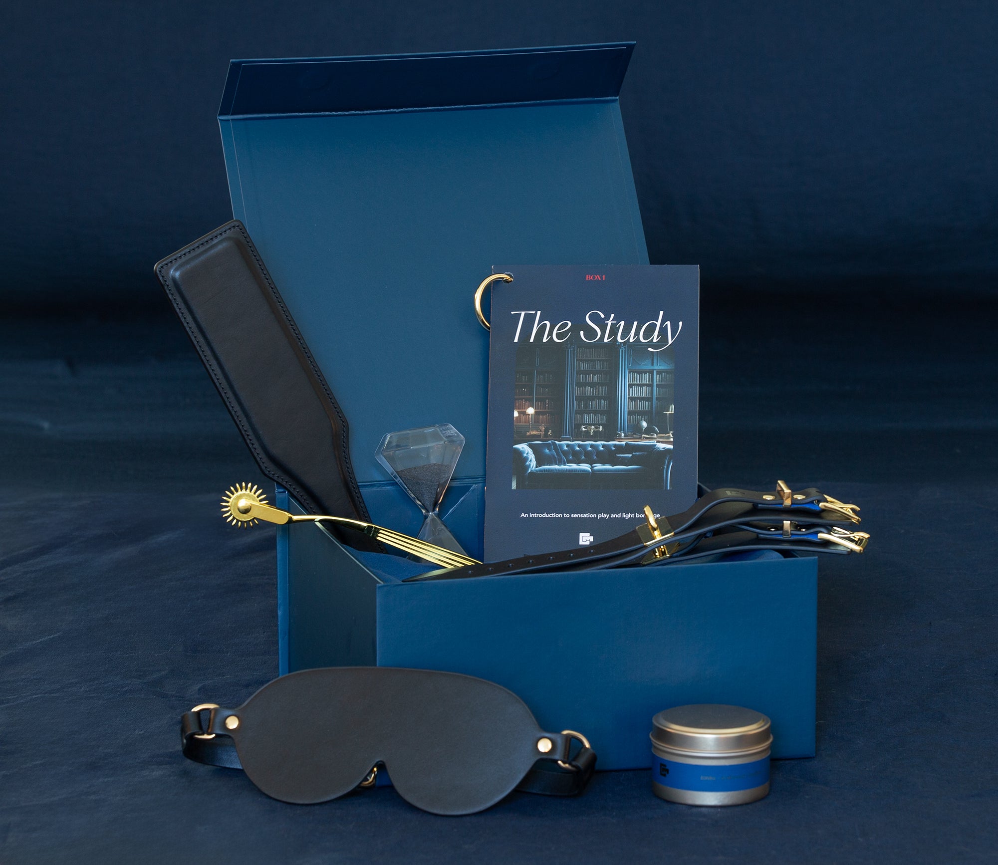 Sex toys displayed in a luxury gift box. Italian leather handcuffs, Italian leather paddle, leather eye mask, massage candle, wartenberg wheel, hourglass, booklet.